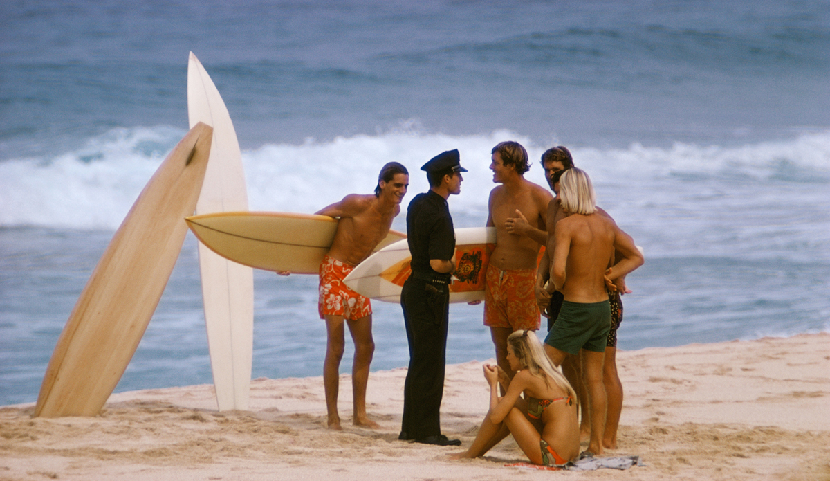 Larry Strada “Wolfman”, Bob Stay, Sherry Haley Juan Shelton and Joey Cabell at Pipeline talking with Hawaii 5-0,  1969. Image: <a href=\"https://www.artbrewer.com/\" target=\"_blank\">Brewer</a>