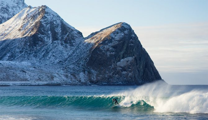 a surfer catches a wave with a snowy mountain behind