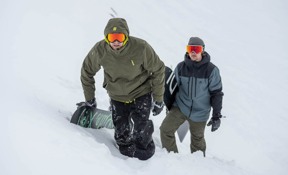 Two snowboarders climb a snowy hillside, one wearing a forum jacket and the other wearing a jones mtn surf jacket.