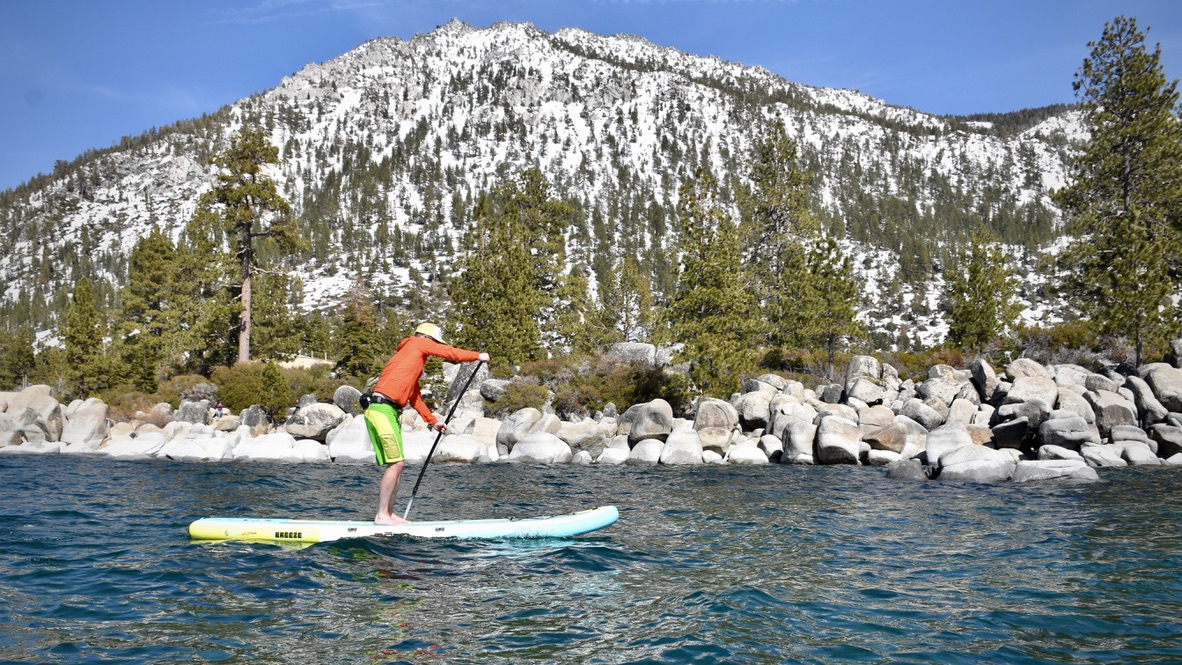 a man testing the bote breeze aero inflatable paddle board on lake tahoe with boulders, trees, and mountains in the background