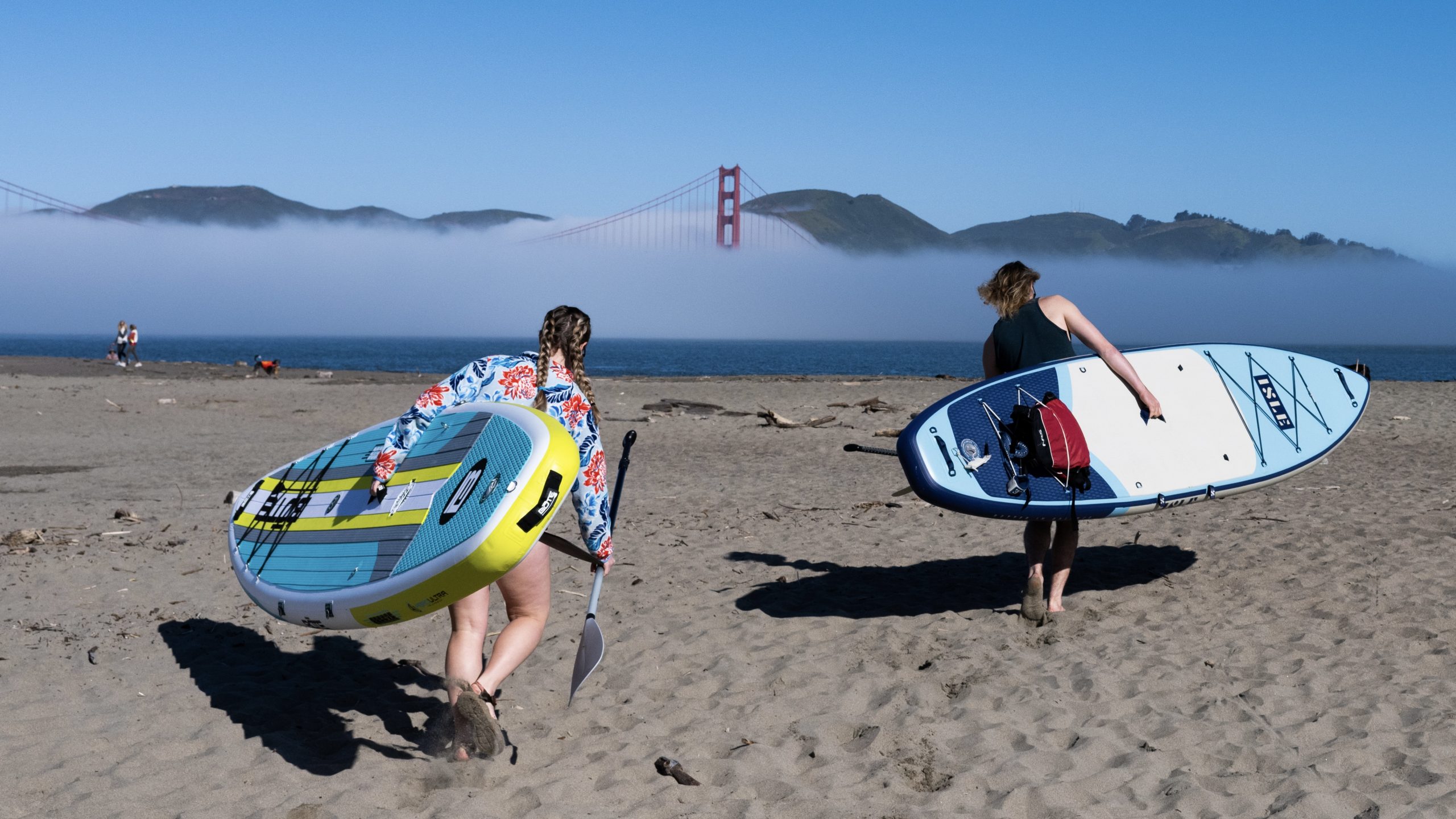 two testers carrying inflatable paddles boards towards San Francisco Bay with golden gate bridge in the background