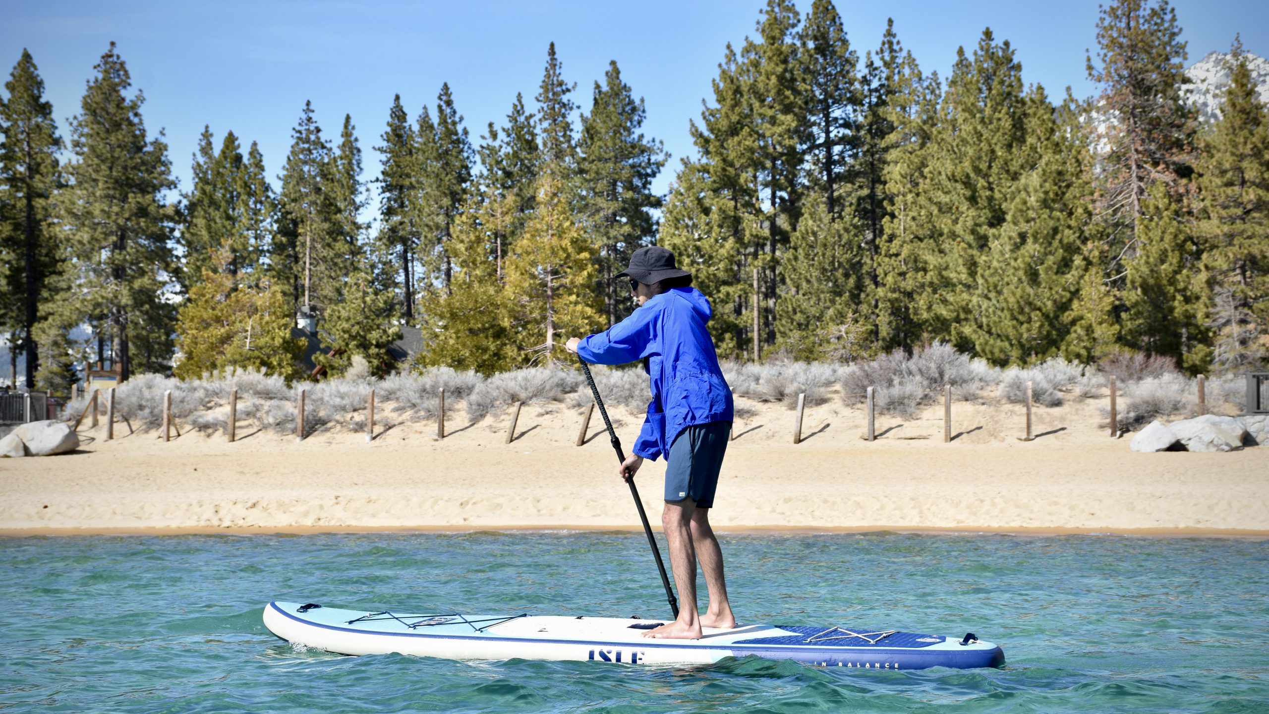a man testing the isle explorer 2.0 inflatable paddle board on lake tahoe with the beach and trees in the background