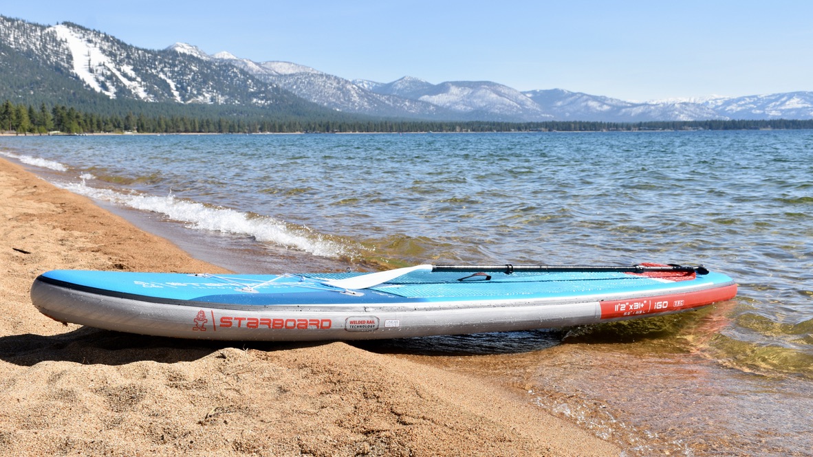 the starboard igo zen inflatable paddle board sitting on the shore of lake tahoe with mountains in the background