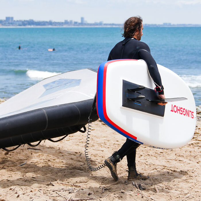 a man carrying the slingshot ltf board from the beach to the water with a wing