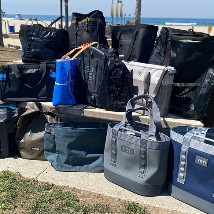 a photo of an assortment of totes, insulated coolers, and backpacks grouped together by the beach. These were the items we tested for our review of the best beach bags.