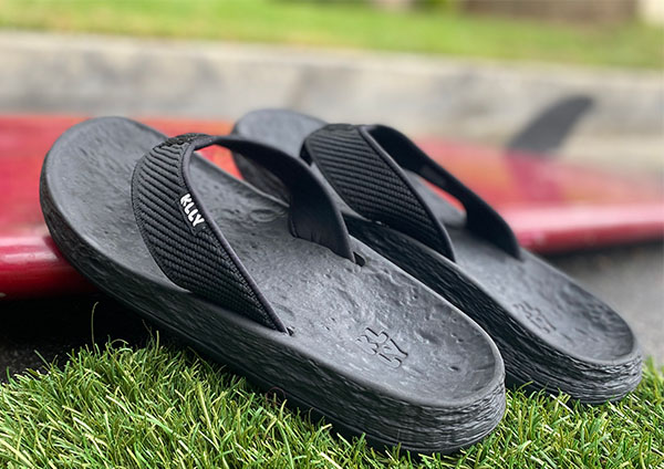 A pair of black flip flops resting on a surfboard. The flip flops are from KLLY which won our pick for best eco friendly sandals.