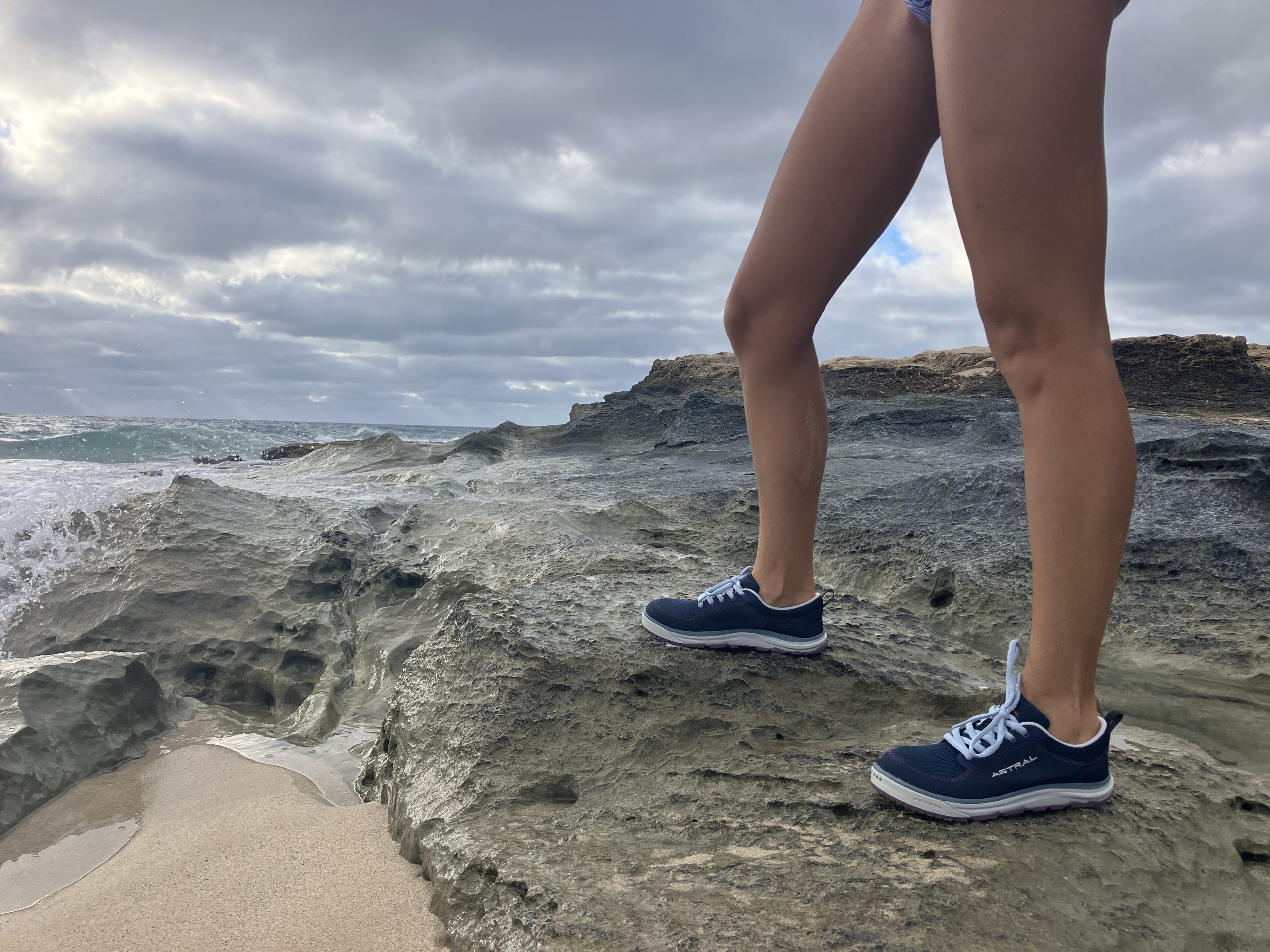 Two legs wearing astral water shoes on a rocky shoreline with the beach in the background