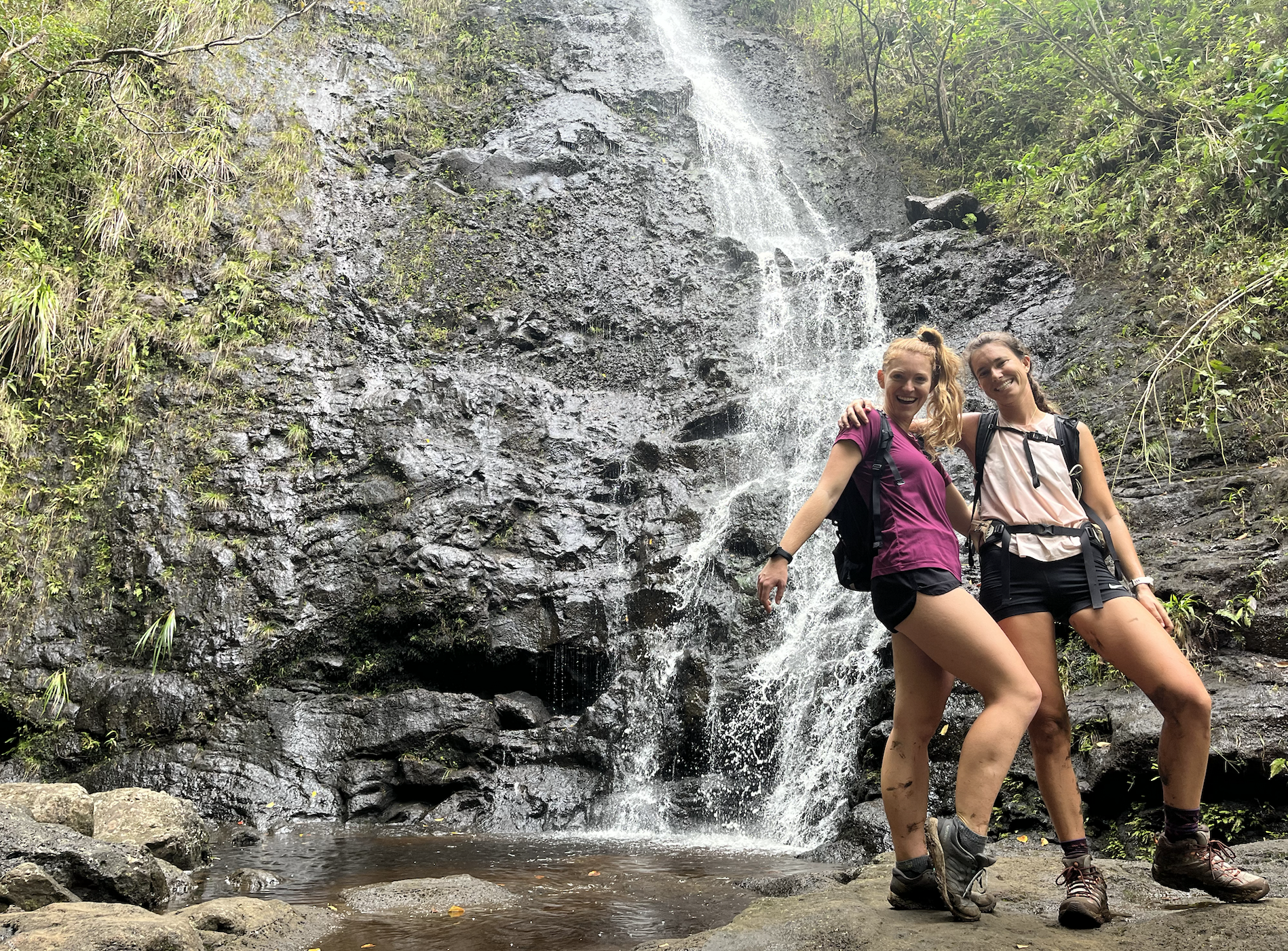two women pose near a waterfall while wearing hiking boots