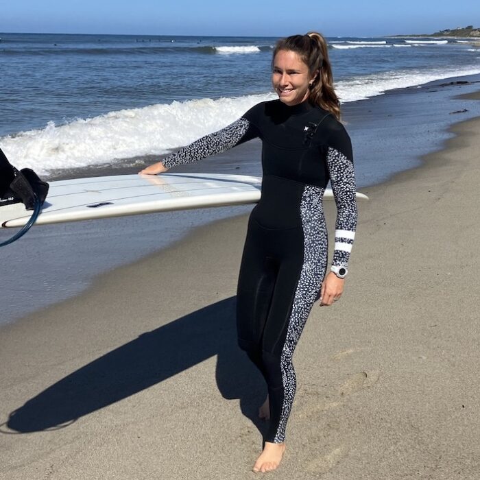 a woman wearing a hurley wetsuit walking on the beach with a surfboard