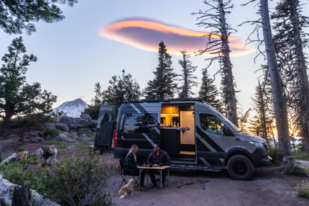 A couple and their dog sit in front of a camper van parked in a wooded area during the sunest.