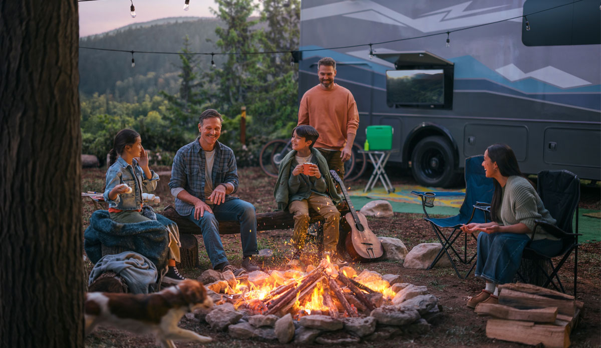 A family sits around a campfire next to their RV in the evening