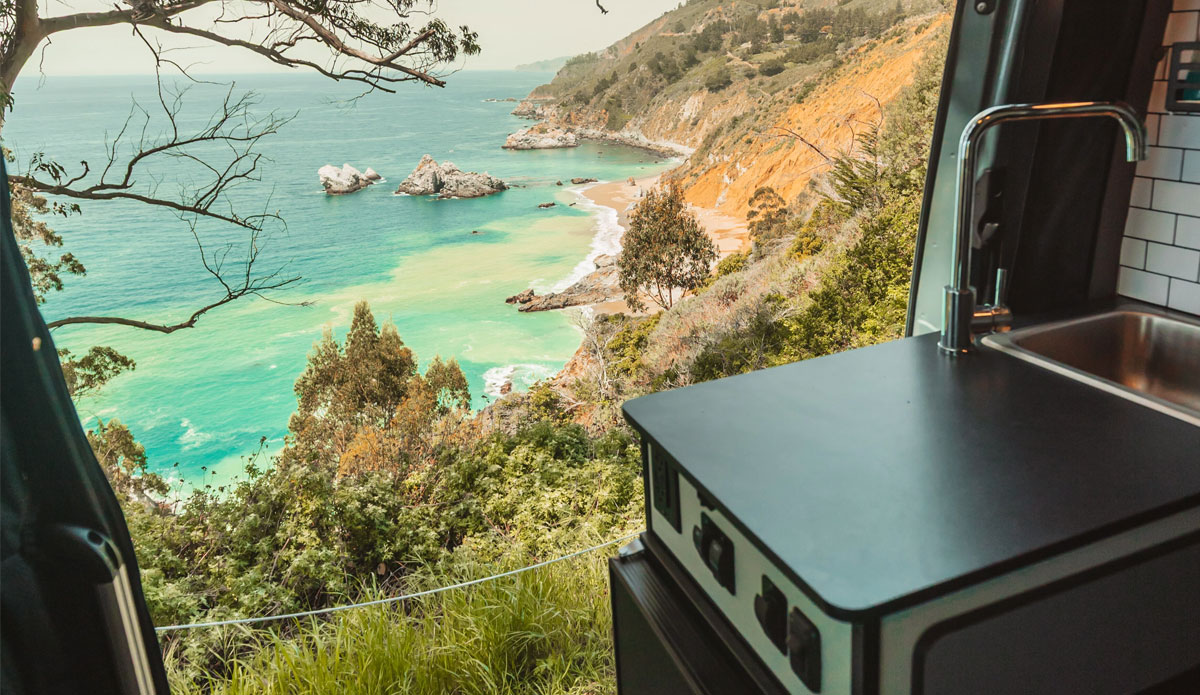 A view of a ocean cliff side overlooking aquamarine waters through the kitchen door of a camper van