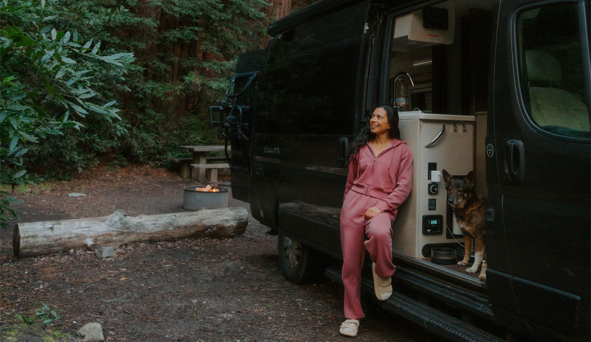 A woman in a pink outfit leans against the open door of her camper van alongside her dog