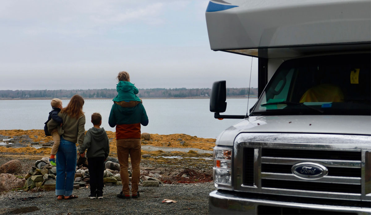 A young family stands outside of their RV facing away from the camera while they look out at a lake