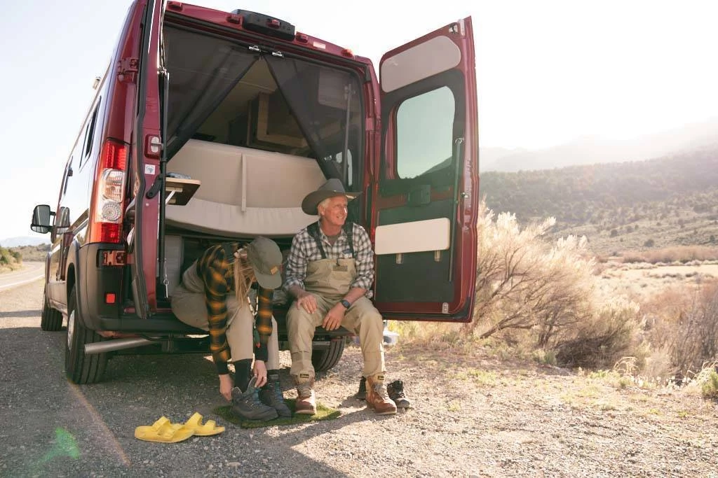 A photo of an older man and younger woman putting on boots to go fly-fishing while seated in the back of their camper van