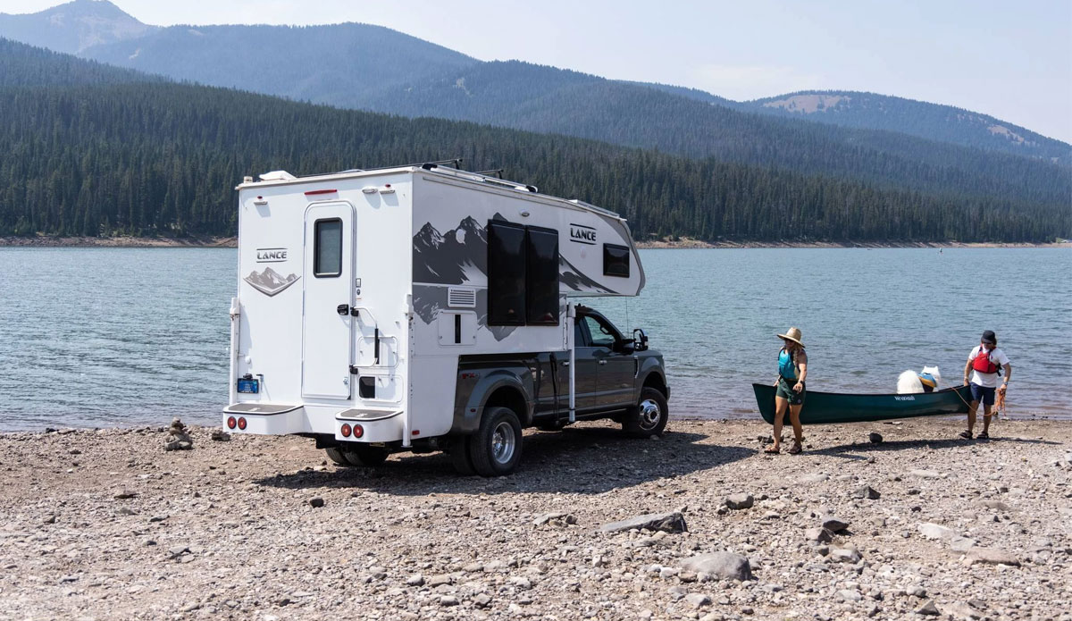 A view of a truck camper and people carrying a canoe towards a lake in the mountains