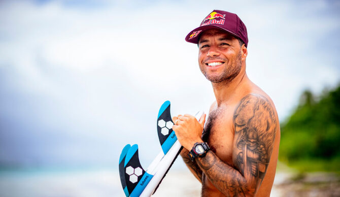 Adriano de Souza smiling with a Red Bull hat while leaning on a surfboard